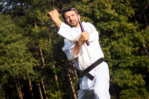 Мастер боевых искусств в белом кимоно практикуется с нунчаку на природе
. - Фото, изображение