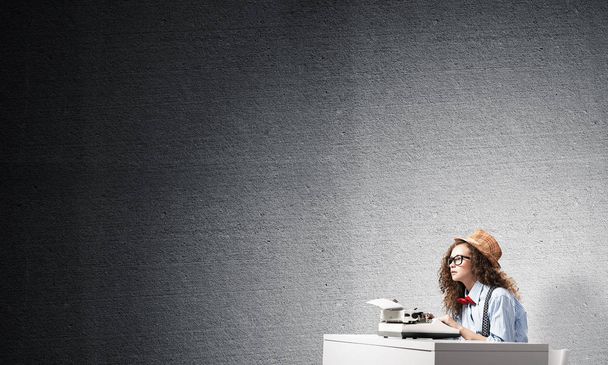 Seitenansicht der jungen schönen Schriftstellerin mit Hut und Brille mit Schreibmaschine, während sie am Tisch vor grauer Betonwand im Hintergrund sitzt. - Foto, Bild