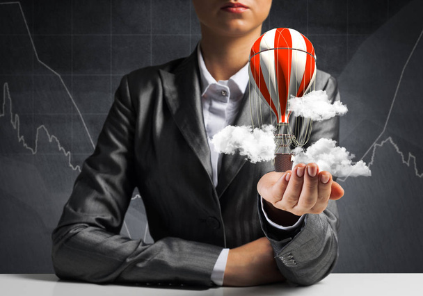 Closeup of business woman in suit presenting flying aerostate among clouds in her palm with business sketches on background. 3D rendering. - Photo, Image