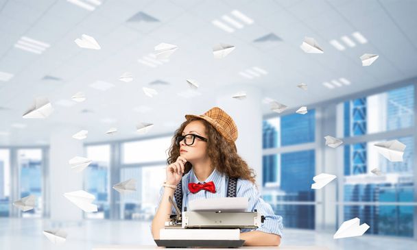 Young woman writer in hat and eyeglasses using typing machine while sitting at the table indoors among flying paper planes and with office view on background. - Foto, afbeelding