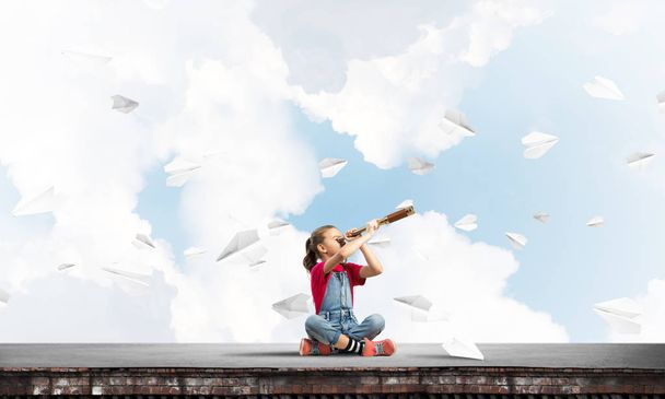 Cute kid girl sitting on house roof and looking in spyglass - Photo, image