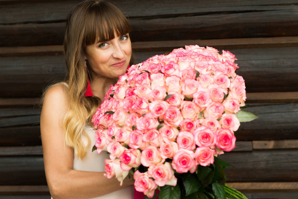 young pretty with a light and long hair girl smiling pleasantly holds a large bouquet of white and red roses standing next to a wooden wall - Photo, Image