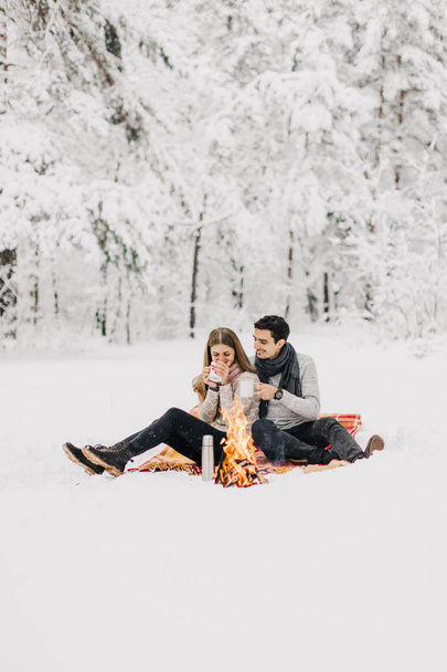 Young couple on a date in the winter forest is heated by the fire and drink cocoa - Фото, изображение