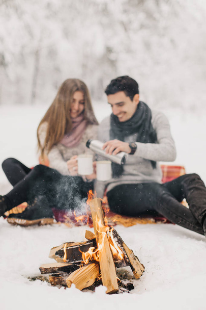 Young couple on a date in the winter forest is heated by the fire and drink cocoa - 写真・画像