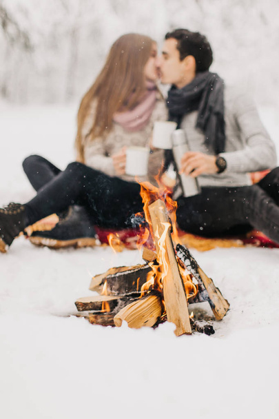 Young couple on a date in the winter forest is heated by the fire and drink cocoa - Fotó, kép