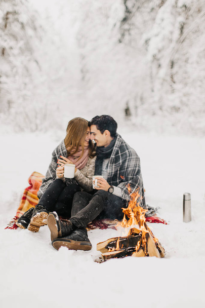 Young couple on a date in the winter forest is heated by the fire and drink cocoa - Fotografie, Obrázek