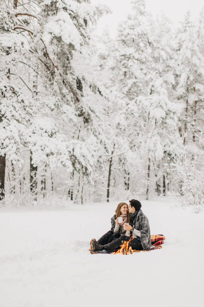 Young couple on a date in the winter forest is heated by the fire and drink cocoa - Fotoğraf, Görsel