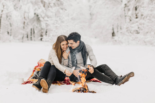 Young couple on a date in the winter forest is heated by the fire and drink cocoa - Photo, Image