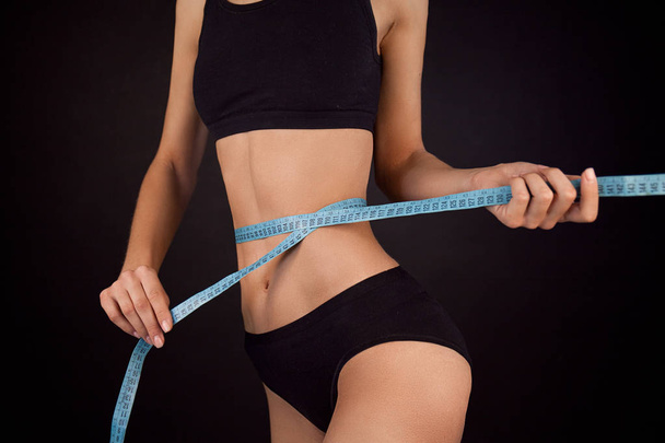 Young Sporty Fitness Girl Wearing Black Sport Underwear Measuring Waist Size With Blue Roulette Black Background - Fotoğraf, Görsel