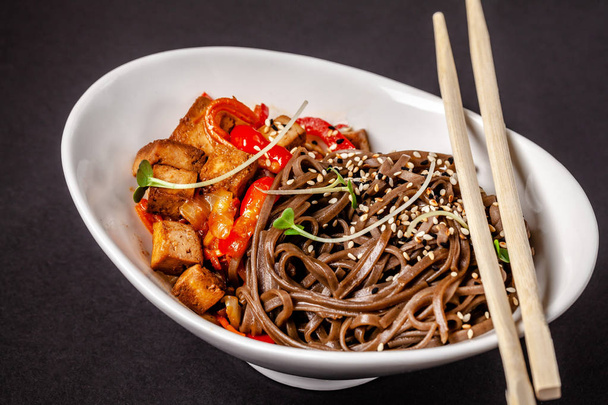 Concept of Asian cuisine. Buckwheat noodles with vegetables and tofu. Carrots, bell peppers, white and black sesame. Japanese or Chinese dish. - Фото, изображение