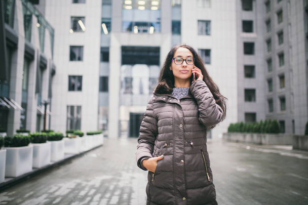 Theme is the business situation. Beautiful young woman of European ethnicity with long brunette hair wearing glasses and coat stands on background of business center and uses phone in hand near ear. - Photo, Image