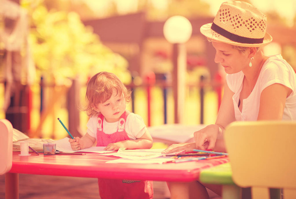 young mother and her little daughter cheerfully spending time together using pencil crayons while drawing a colorful pictures in the outside playschool - Foto, Imagem