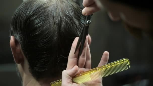 Guy with a beard and a dark hair in the illuminated barbershop. Barber is cutting his hair with scissors and an yellow comb. Closeup video recording. - Filmagem, Vídeo