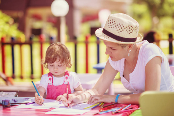 young mother and her little daughter cheerfully spending time together using pencil crayons while drawing a colorful pictures in the outside playschool - Photo, image