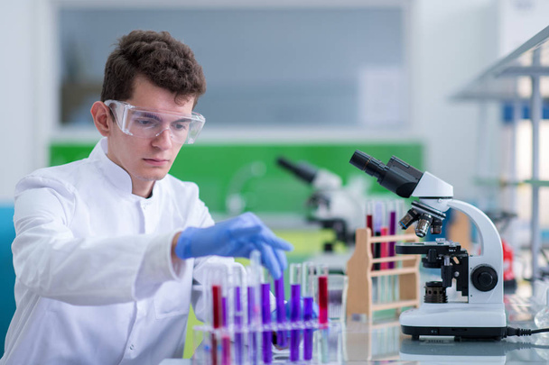молодой студент в защитных очках проводит химический эксперимент в яркой лаборатории, образовании и медицине - Фото, изображение