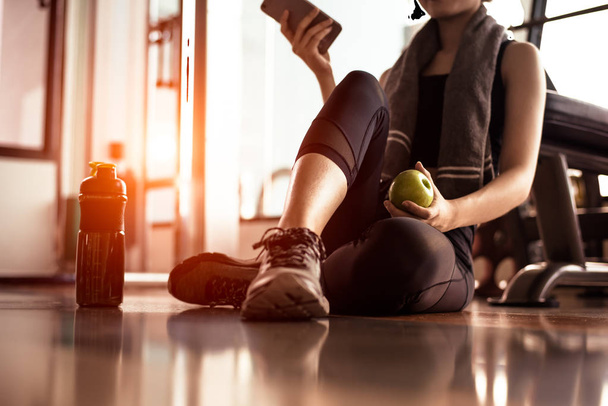 Close up of woman using smart phone and holding apple while workout in fitness gym. Sport and Technology concept. Lifestyles and Healthcare theme. - Photo, Image