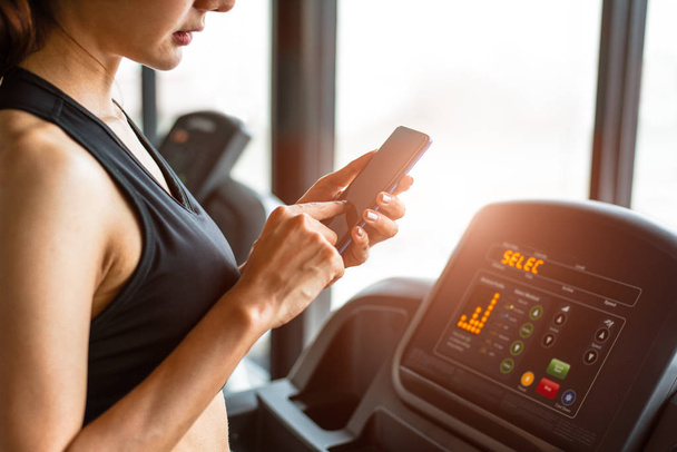 Woman using smart phone when workout or strength training at fitness gym on treadmill. Relax and Technology concept. Sports Exercise and Health care theme. Happy people and Comfortable application - Photo, Image