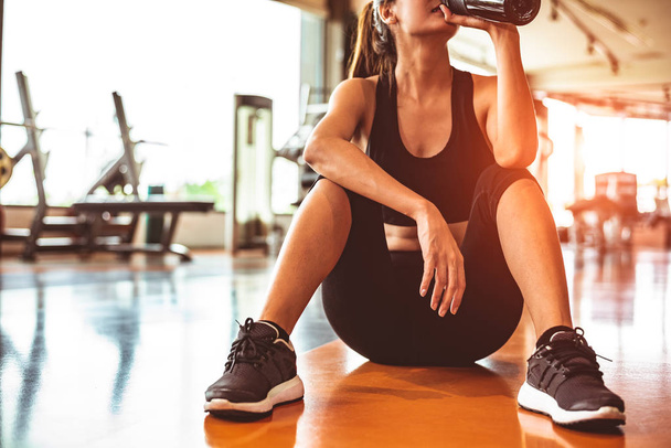 Спортивная женщина отдыхает после тренировки или тренировки в тренажерном зале. Сидеть и пить протеиновый коктейль или питьевую воду на полу. Силовые тренировки и бодибилдер мышечной теме. Теплый и прохладный тон
  - Фото, изображение