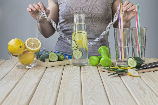 Woman measures her waist at home in  kitchen. Detox drink from cucumbers and lemons, dumbbells for fitness. Concept: healthy lifestyle - Photo, Image