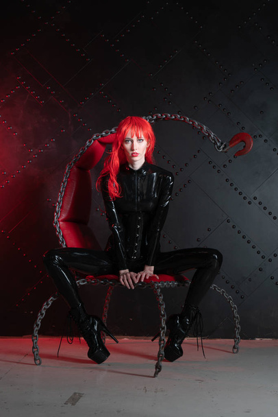 dangerous cruel fetish girl with red hair posing in black bdsm latex clothes at the chair from the chains in the style of sadomasochism - Foto, imagen