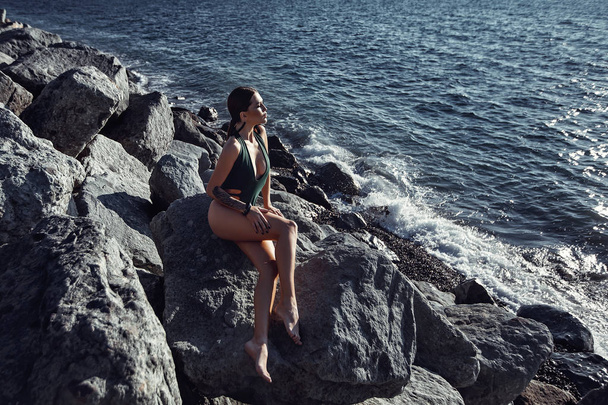 Hermosa chica en traje de baño sentada posando en una roca junto al océano. Isla, puesta del sol, descanso, sol, viajes, libertad
. - Foto, Imagen