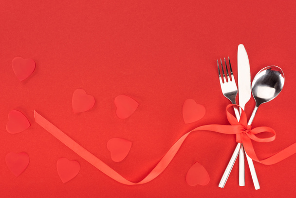 elevated view of cutlery wrapped by festive ribbon near heart symbols isolated on red, st valentine day concept - Photo, Image