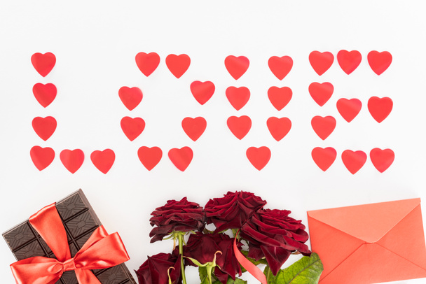 top view of lettering love made of heart symbols, chocolate wrapped by festive ribbon, red roses and envelope isolated on white, st valentine day concept - Photo, Image