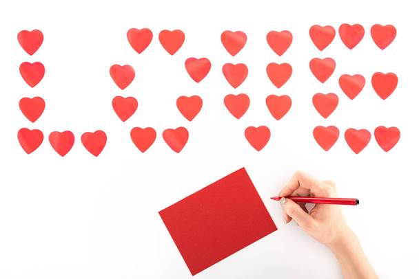 cropped image of woman writing greeting card under lettering love made of red heart symbols isolated on white, st valentine day concept - Foto, afbeelding