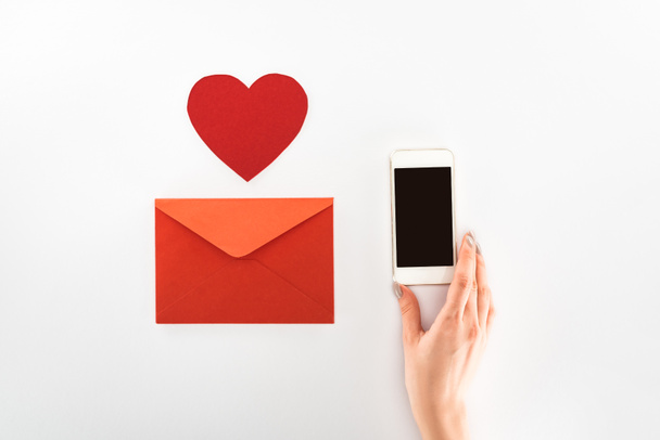 cropped image of woman holding smartphone with blank screen near heart symbol and red envelope isolated on white, st valentine day concept - Photo, Image