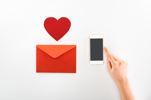 abgeschnittenes Bild einer Frau mit Smartphone mit leerem Bildschirm in der Nähe des Herzsymbols und rotem Umschlag auf weißem Papier, Valentinstag-Konzept - Foto, Bild