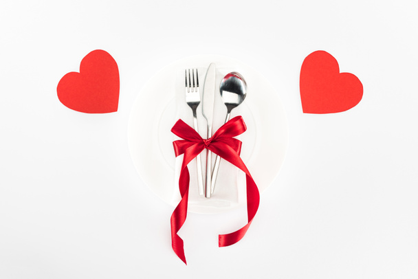 vista elevata di piatto con posate avvolto da fiocco nastro festivo vicino ai simboli del cuore isolato su bianco, concetto di San Valentino
 - Foto, immagini