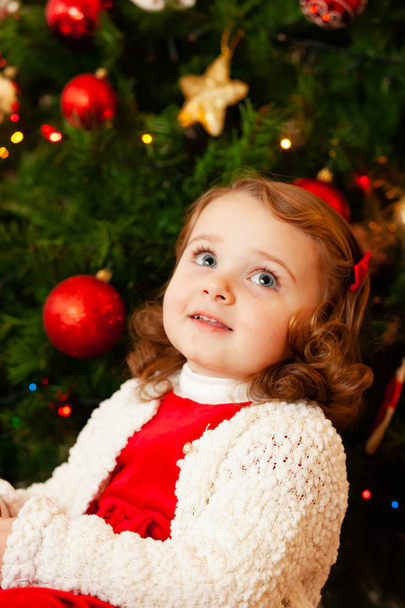 Portrait of a beautiful little child with red dress near Christmas tree. - Фото, зображення
