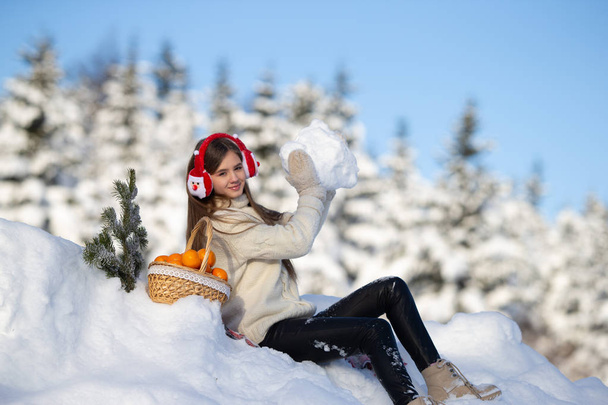 A young girl walks in the winter forest with a basket of mandarin in headphones Santa Claus. - Photo, Image