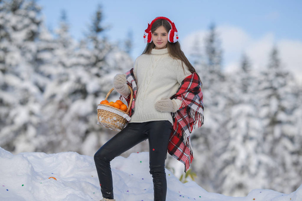 A young girl walks in the winter forest with a basket of mandarin in headphones Santa Claus. - Photo, Image