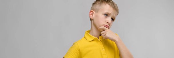 Portrait of cute little blonde caucasian boy in yellow t-shirt unsure puzzled on gray background - Photo, Image