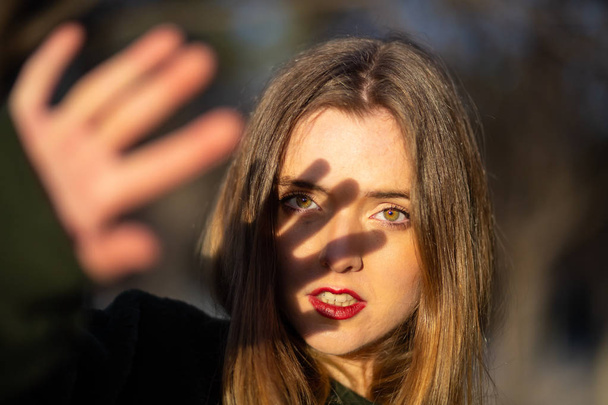 Lovely young female with bright makeup trying to cover face with hand from sunlight while standing on blurred background of park - Photo, Image