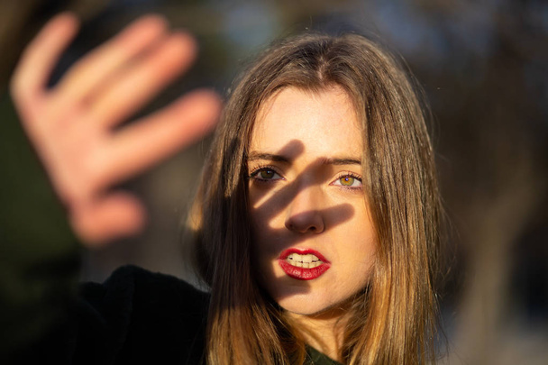 Preciosa joven hembra con maquillaje brillante tratando de cubrir la cara con la mano de la luz del sol mientras está de pie sobre el fondo borroso del parque
 - Foto, Imagen