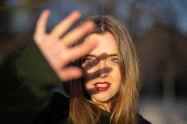 Lovely young female with bright makeup trying to cover face with hand from sunlight while standing on blurred background of park - Photo, Image