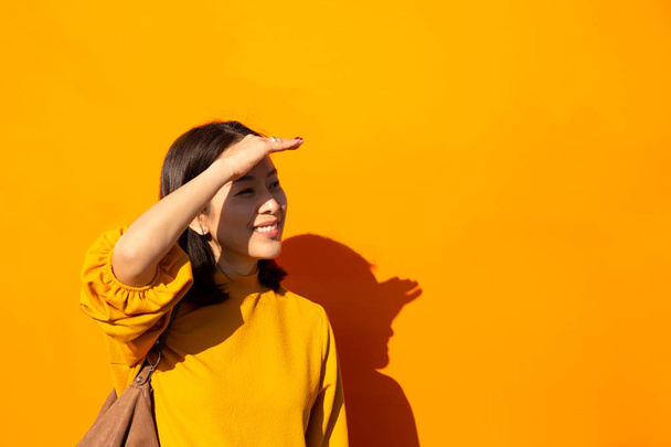 Ázsiai nő, mosolygós narancs felső kezét a homlokán látszó-on fényképezőgép, narancssárga háttérben - Fotó, kép