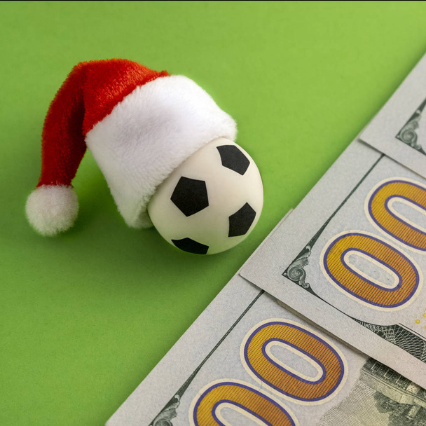 New Year's soccer ball in a red Santa Claus hat next to the hundred dollar bills on a green background. Square frame for instagram. The concept of sports betting. Money, christmas and football. - Photo, Image