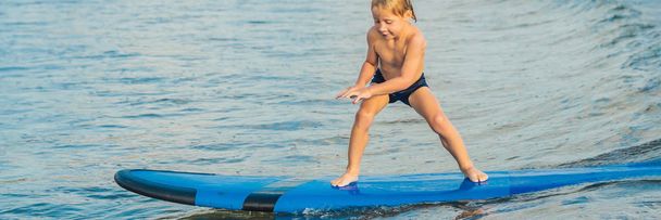 Pieni poika surffaamassa trooppisella rannalla. Lapsi surffilaudalla meren aallolla. Aktiivinen vesiurheilu lapsille. Lapsi ui surffaten. Surffaustunti lapsille BANNER, LONG FORMAT
 - Valokuva, kuva