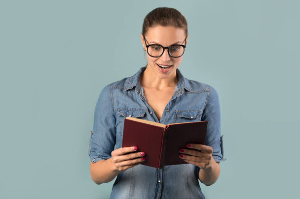 beautiful young girl in glasses holding a book with astonishment on her face on a blue background - Photo, image
