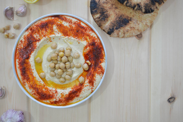 plato de hummus casero tradicional sobre fondo rústico de mesa con pimentón rojo aceite de oliva, ajo, limón, tahini y pan de pita Imagen
 - Foto, imagen