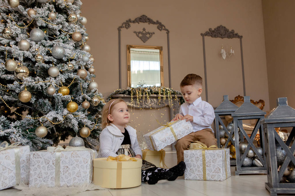 Cute Girl and boy opening Xmas presents. Children under Christmas tree with gift boxes. Decorated living room with traditional fire place. Cozy warm winter evening at home. - Foto, Bild