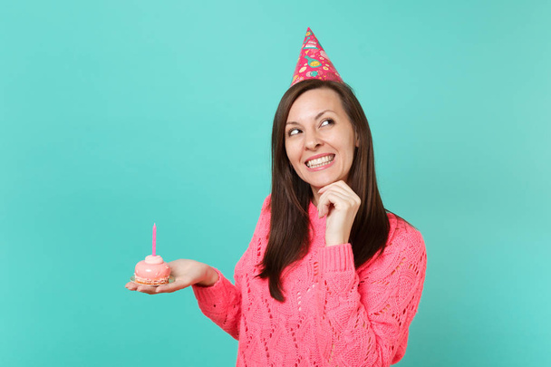 verträumte junge Frau in gestricktem rosa Pullover, Geburtstagsmütze nach oben, Handstütze am Kinn, Handtorte mit Kerze vor blauem Hintergrund. Menschen Lebensstil Konzept. Attrappe Kopierraum - Foto, Bild