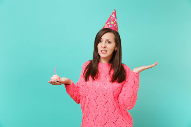 Озадаченная молодая женщина в вязаном розовом свитере, шляпа на день рождения, указывающая на руку, держащая торт со свечой, выделенной на фоне синей стены, студийный портрет. Идея образа жизни людей. Пространство для копирования
 - Фото, изображение