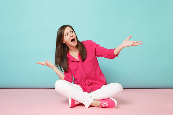Volledige lengte portret van boze jonge vrouw in roze overhemd blouse, witte broek, zittend op de vloer geïsoleerd op helder roze pastel blauwe muur achtergrond studio. Fashion lifestyle concept. Bespotten kopie databaseruimte - Foto, afbeelding