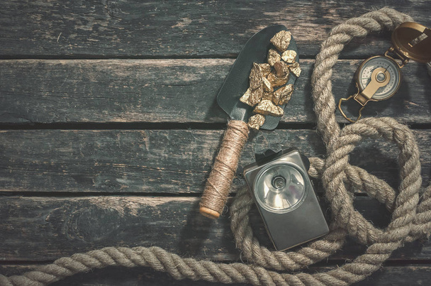 Compas, gold nuggets on the spade, rope and vintage flashlight on the aged wooden table background with copy space. Treasure hunter or goldminer concept. - Valokuva, kuva