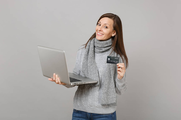 Mujer bastante joven en suéter gris, bufanda que trabaja en el ordenador portátil PC, la celebración de la tarjeta bancaria de crédito aislado en fondo gris de la pared. Estilo de vida saludable, consultoría de tratamiento en línea, concepto de temporada fría
 - Foto, Imagen
