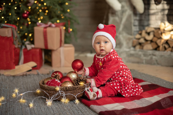 kleines Mädchen mit Weihnachtsmütze und Weihnachtsdekoration am Baum sitzend, Neujahrskonzept  - Foto, Bild